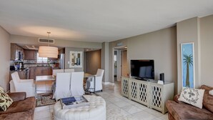 A modern living room with a dining area, kitchen, and a TV on a cabinet. - Station One - 8I From - Oceanfront condo with community pool, tennis, beach (Wrightsville Beach)