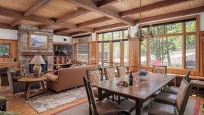 A cozy living room with a fireplace, wooden furniture, and large windows offering a view of the outdoors.