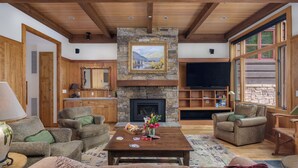 A spacious living room with a stone fireplace, wooden beams, and large windows.