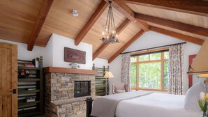 A bedroom with a stone fireplace, a bed with a white comforter, a wooden ceiling, and a large window with curtains.