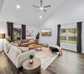 A spacious living room with a sofa, coffee table, and a black bench. There are two windows with curtains, a ceiling fan, and a large painting on the wall.