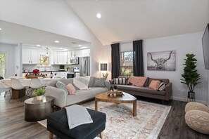 Ein modernes Wohnzimmer mit Sofa, Couchtisch und Hocker, ein Essbereich mit Tisch und Stühlen sowie eine Küche mit weißen Schränken und Geräten.
