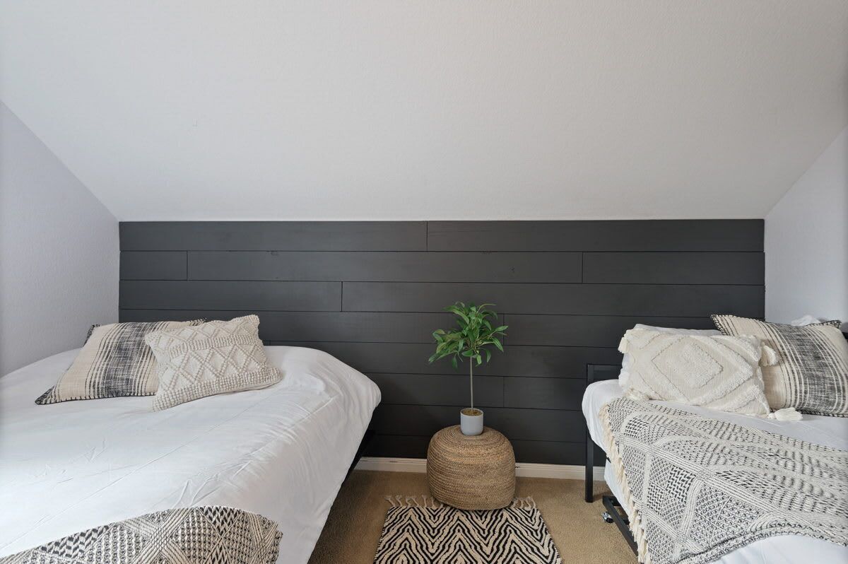 A bedroom with two beds, a dark headboard, a woven pouf, and a potted plant.