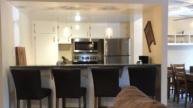 A modern kitchen with white cabinets, a microwave, and a refrigerator. There are three bar stools and a brown sofa in the living area. - Horizons single story ground floor 2 bdrm (Mammoth Lakes)