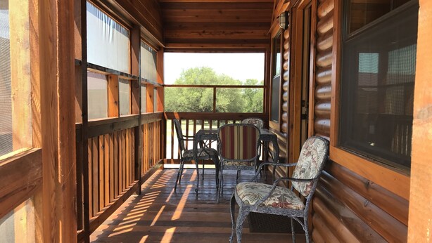 Un porche de madera con barandilla, dos sillas de mimbre y vistas a árboles.