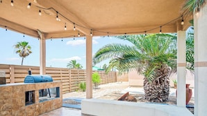 Eine überdachte Terrasse im Freien mit Grill, Sitzgelegenheit und Palmen.