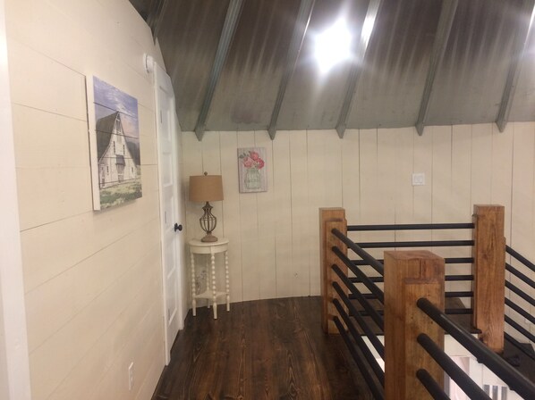 Une pièce avec un plancher en bois, une porte blanche, une petite table avec une lampe, une rambarde en bois et un mur orné d’un tableau et d’une affiche.