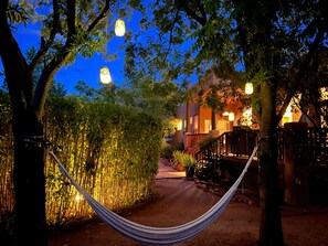 A hammock in a garden path leading to a building with warm lighting and trees.