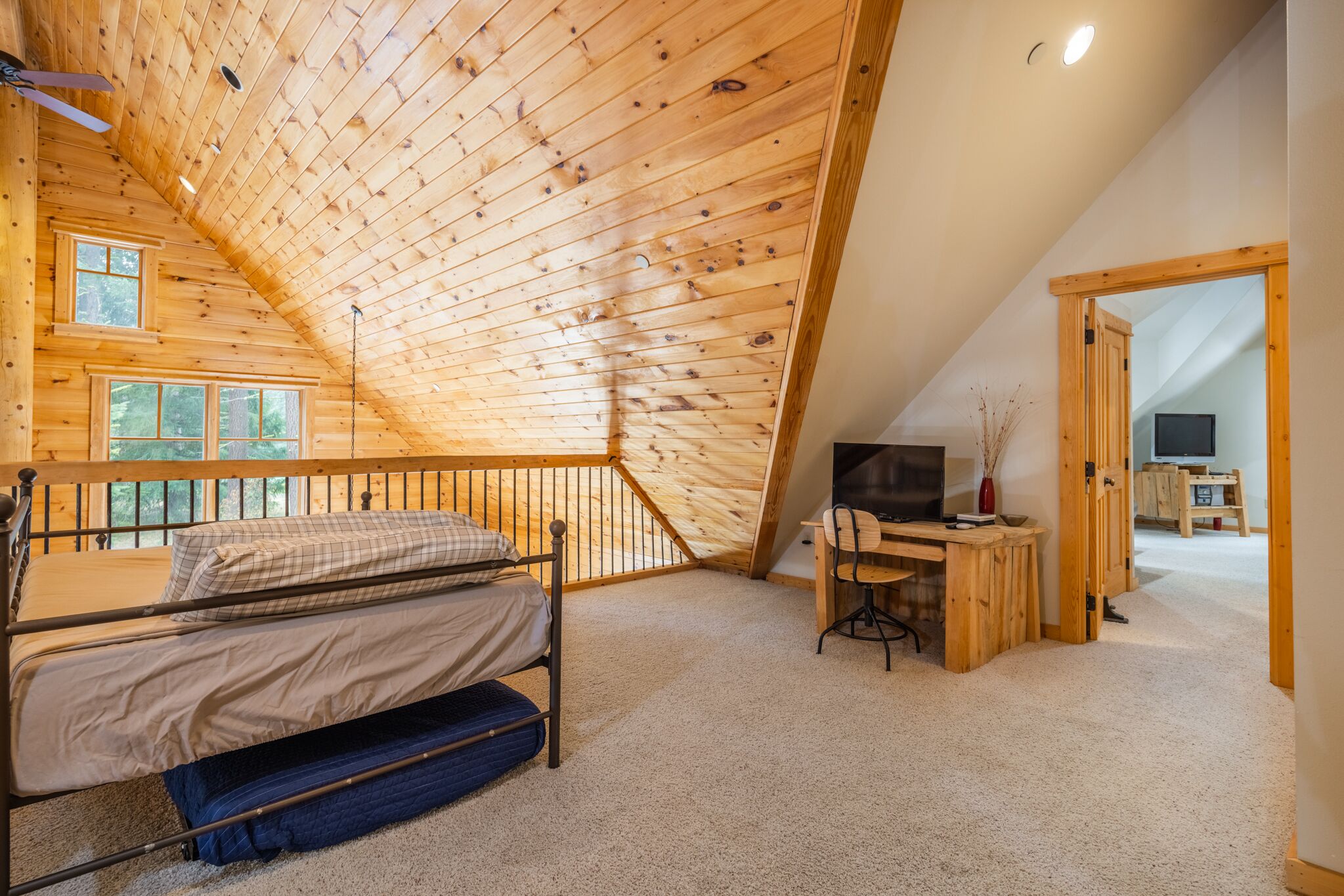 A cozy attic bedroom with a wooden sloped ceiling, a bunk bed, a desk with a chair, and a television.