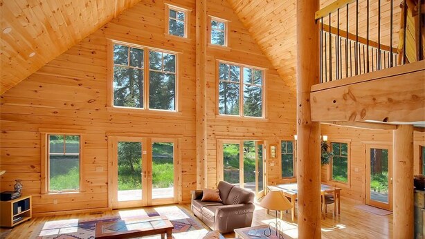 A spacious wooden cabin with large windows, a loft area, and a cozy living room with a sofa and coffee table.
