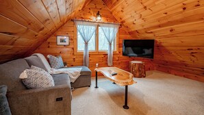 Ein gemütliches Holzhaus mit einer Couch, einem Couchtisch, einem Fernseher und einem Fenster mit Vorhängen.