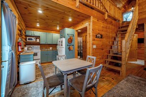 Interior de una cabaña de madera con una mesa de comedor, sillas, una cocina con electrodomésticos y una escalera.