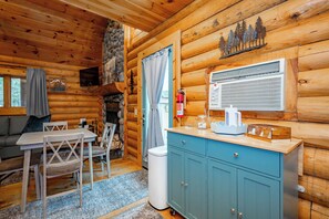 Interior de una cabaña de troncos con zona de comedor, un armario azul y chimenea de piedra.
