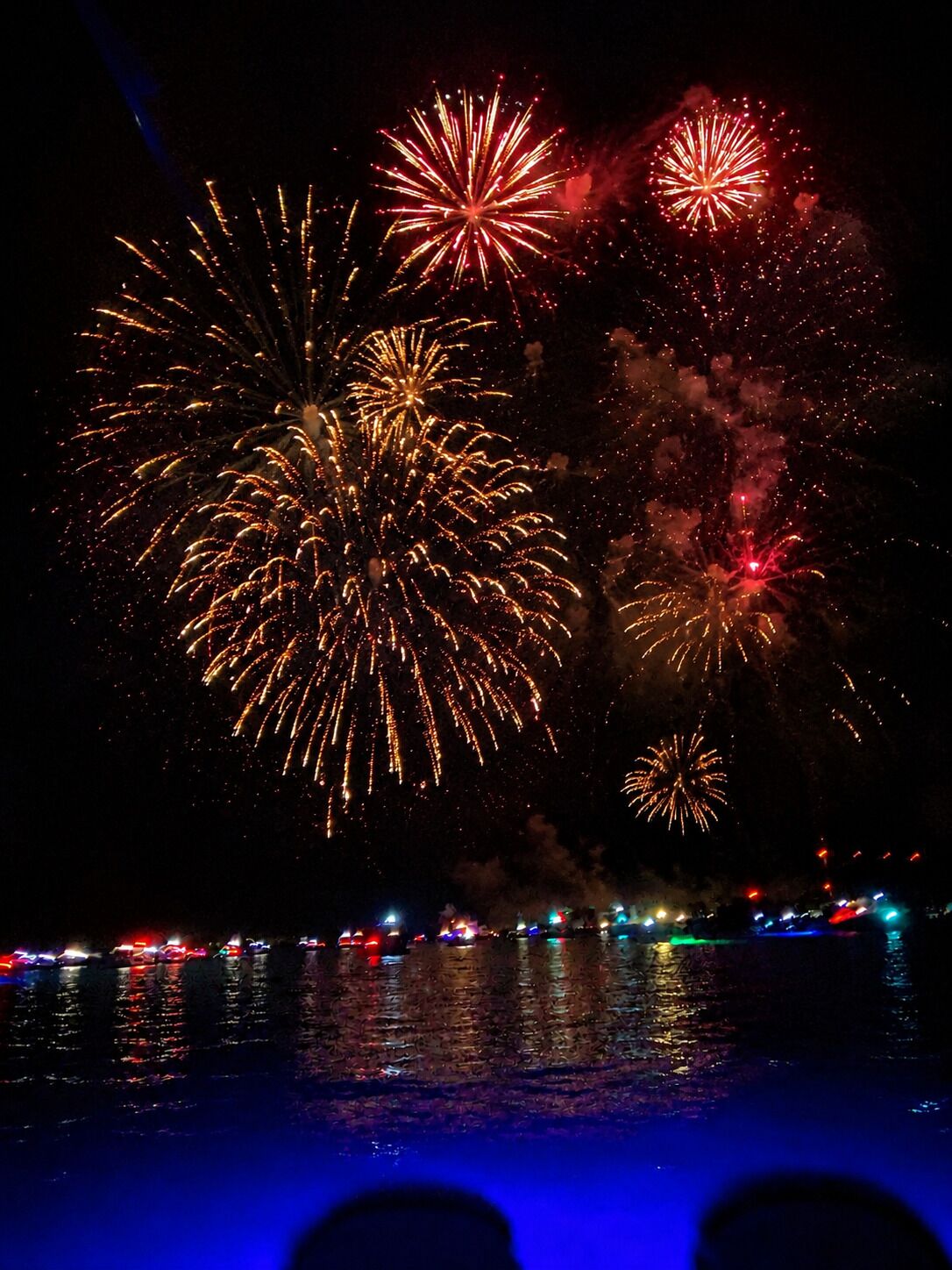 Vista nocturna de fuegos artificiales sobre un cuerpo de agua, con reflejos y un horizonte urbano distante.