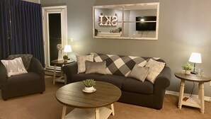 Un salón con un sofá, dos sillones, una mesa de centro y dos mesitas auxiliares con lámparas. Un espejo grande con un televisor está colocado en la pared.