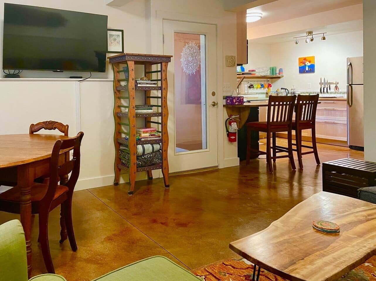 A dining area with a wooden table and chairs, a television mounted on the wall, and a kitchen visible through an open door.