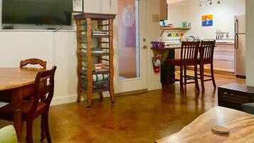 A dining area with a wooden table and chairs, a television mounted on the wall, and a kitchen visible through an open door.