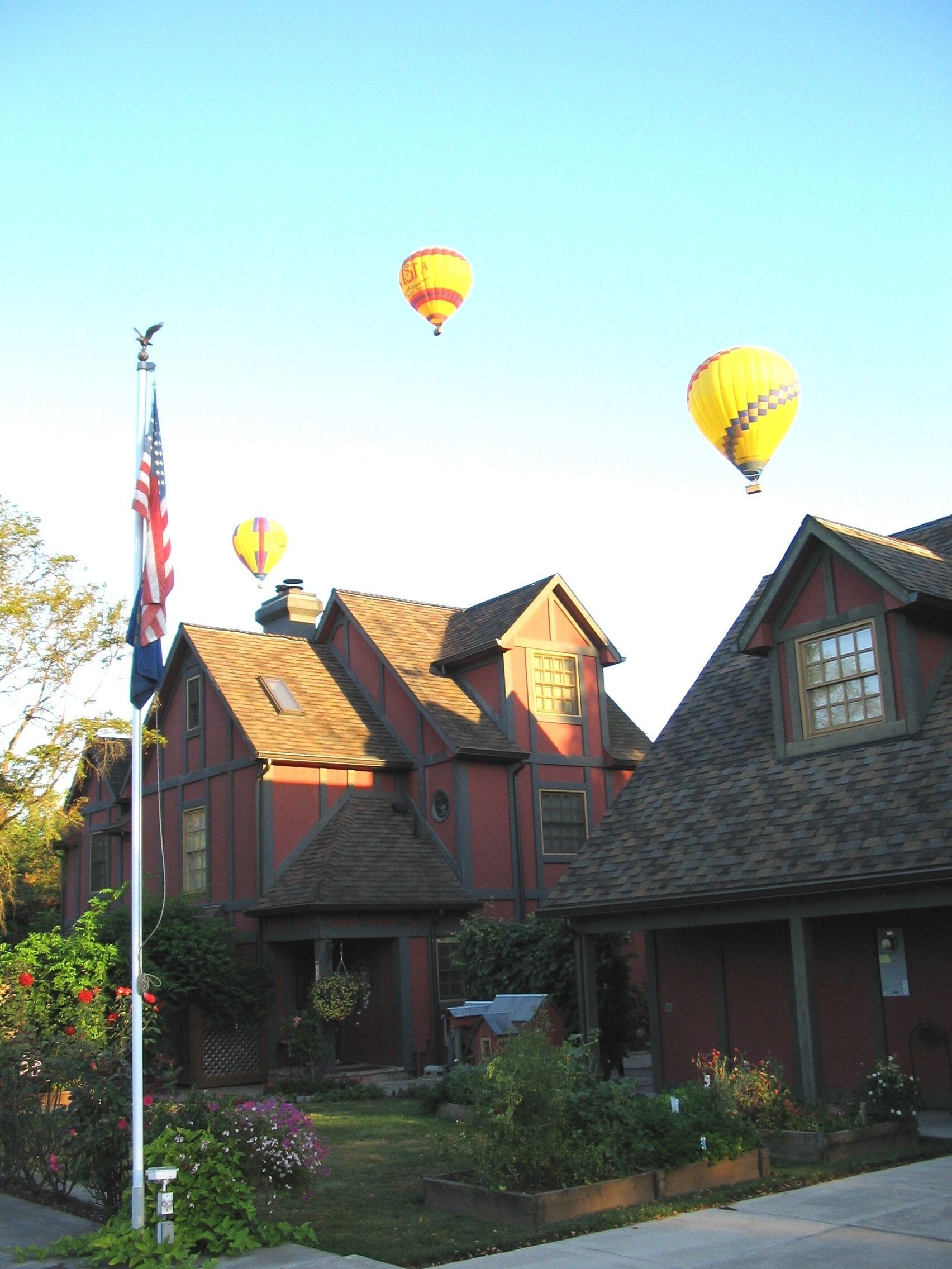 Een klassiek rood huis met een schuine dak, een vlaggenmast met de Amerikaanse vlag, en drie luchtballonnen in de lucht.