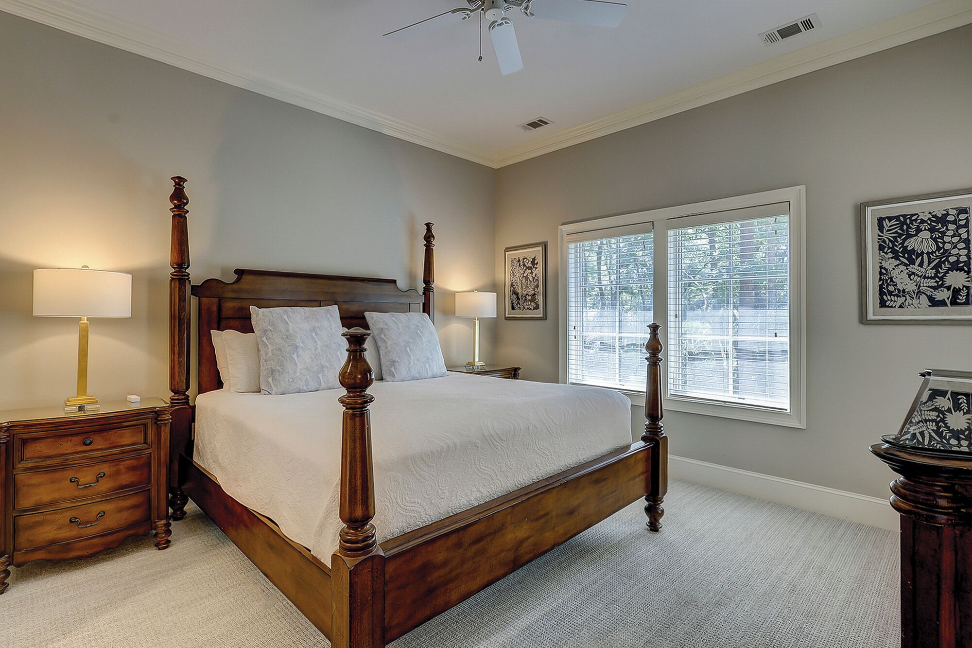A four-poster bed with a wooden frame, two bedside tables with lamps, a large window with blinds, and framed artwork on the walls.