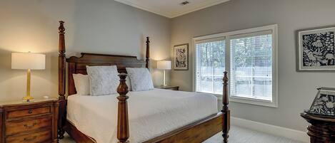 A four-poster bed with a wooden frame, two bedside tables with lamps, a large window with blinds, and framed artwork on the walls.