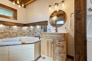 A bathroom with a stone wall, a wooden vanity, a round mirror, and a bathtub. - Canyon View Treehouse- Summer Vacation With A View (Jasper)