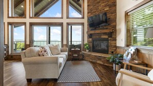 A spacious living room with a fireplace, large windows, and a ceiling fan. - Canyon View Treehouse- Summer Vacation With A View (Jasper)