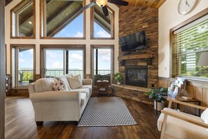 A spacious living room with a fireplace, large windows, and a ceiling fan. - Canyon View Treehouse- Summer Vacation With A View (Jasper)