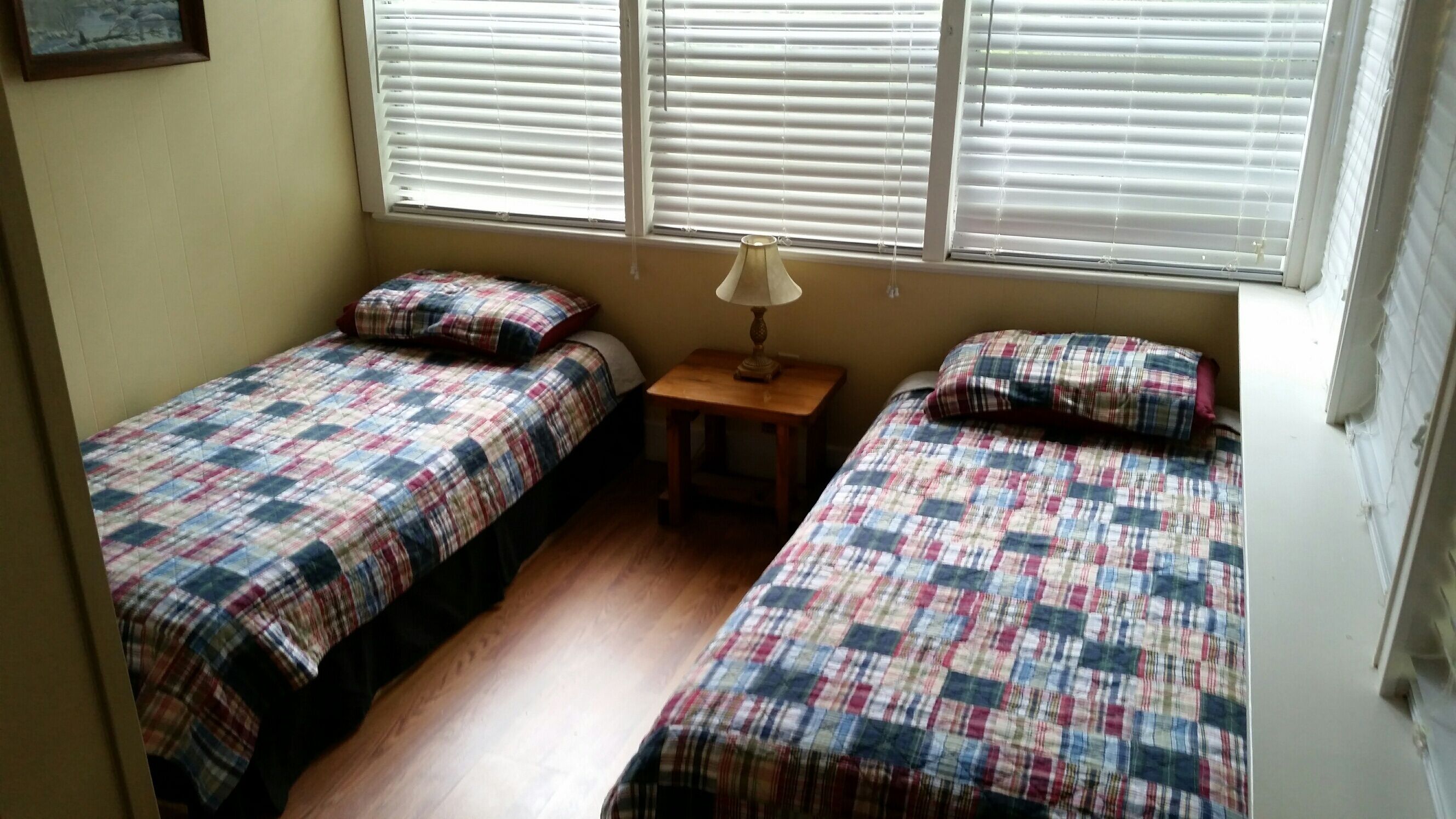 A small, cozy room with two single beds, each with a plaid bedspread and pillows. There is a small wooden nightstand with a lamp between the beds. The room has a window with blinds, and the walls are painted a light color.