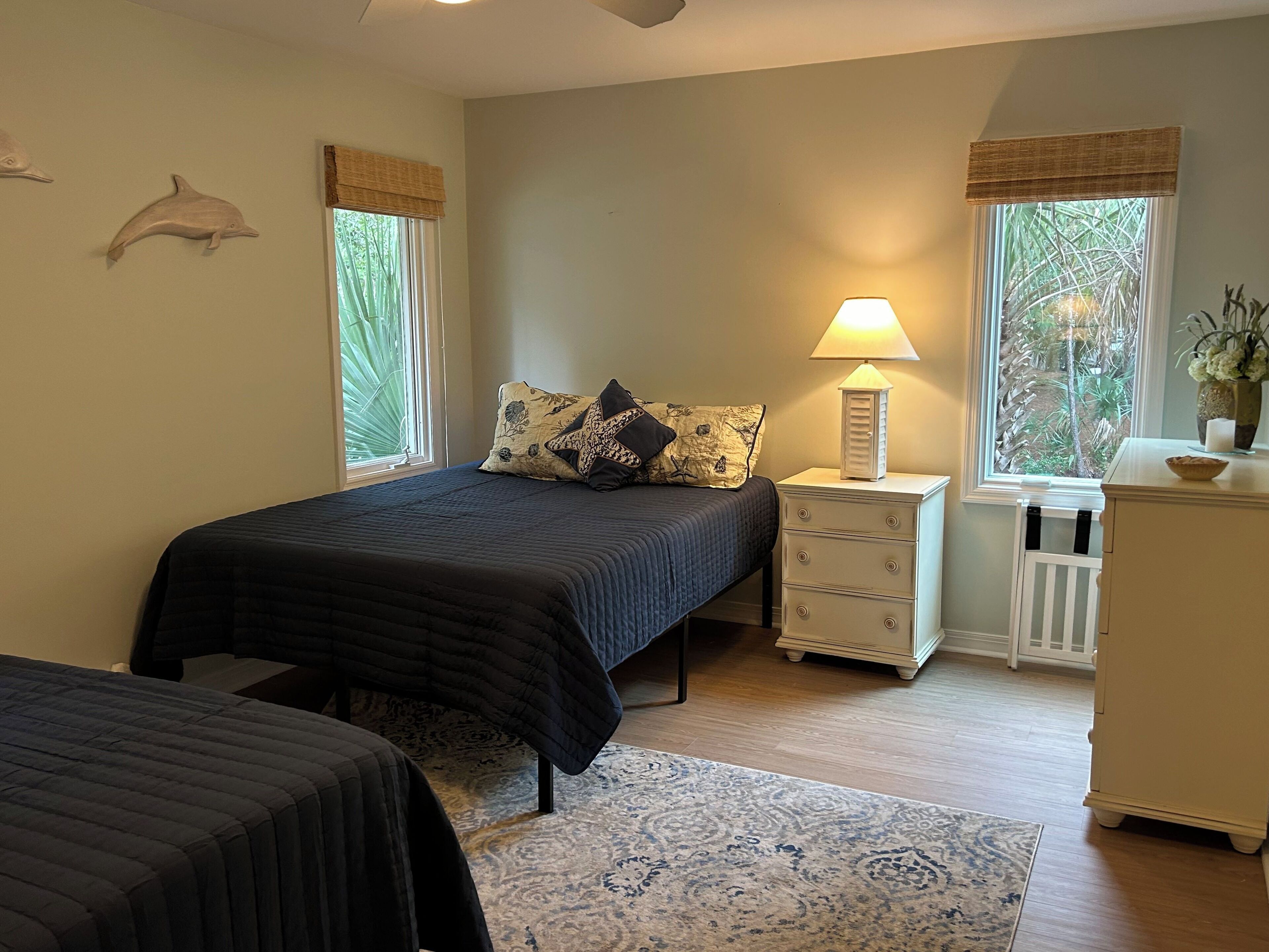 A bedroom with a bed, bedside table, lamp, dresser, and two windows with curtains.