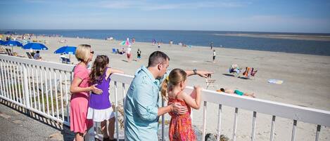 A beach scene with a white railing, people on the beach, and umbrellas.