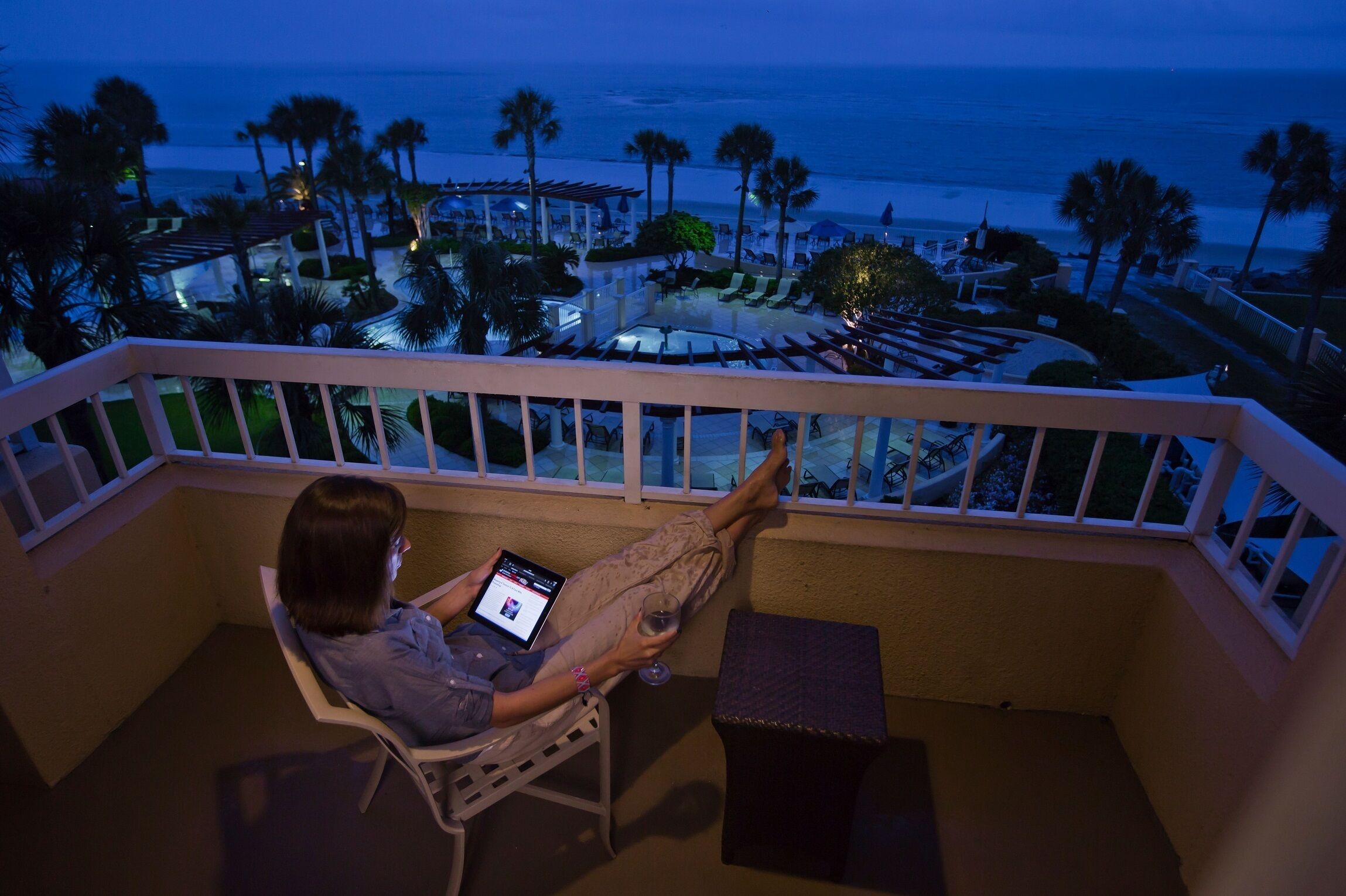 A balcony with a view of the beach, a lounge chair, and a person using a tablet.