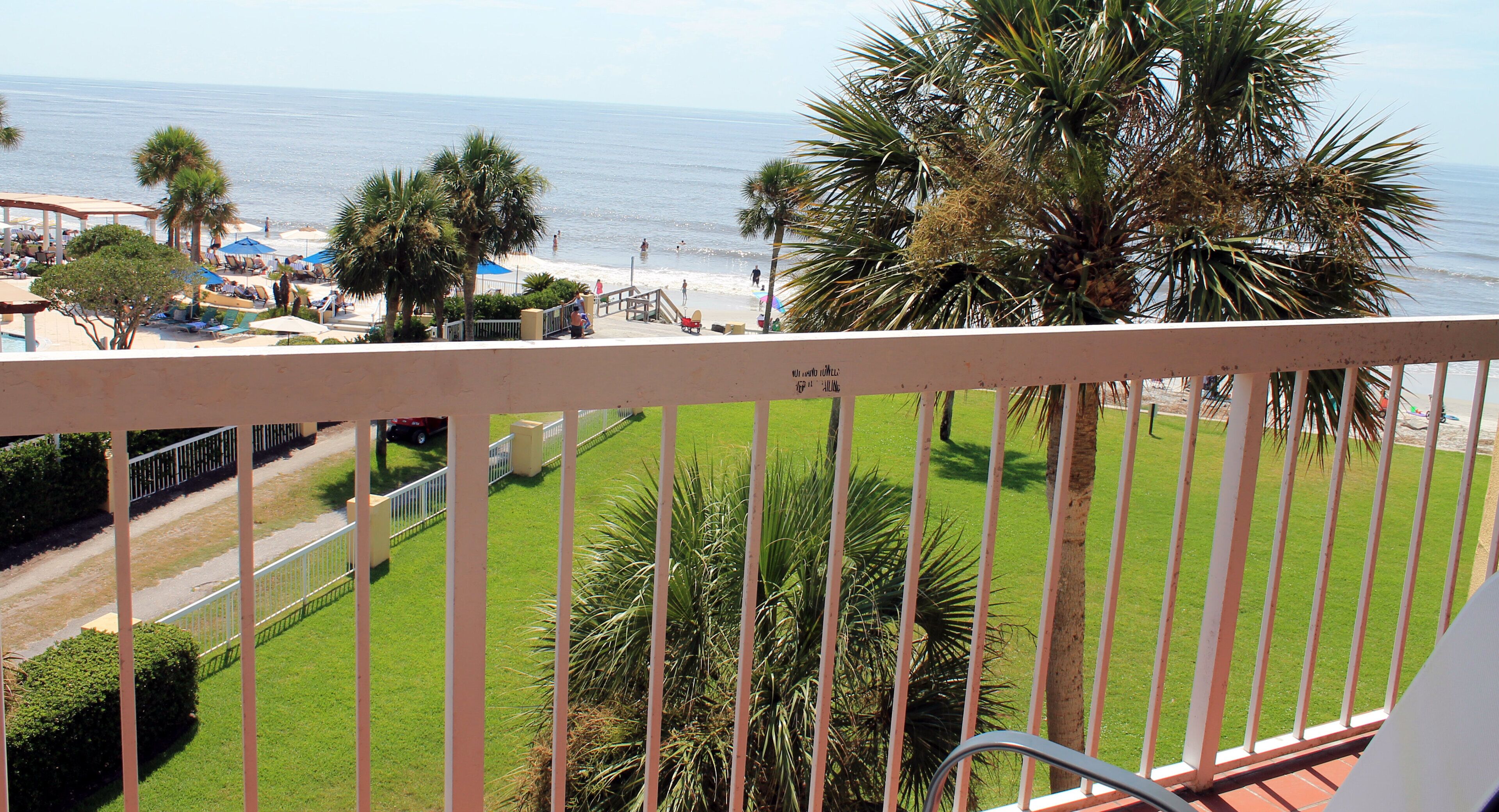 A balcony with a view of the beach, palm trees, and a clear sky.
