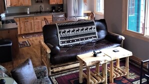 Una sala de estar acogedora con un sofá de cuero, una mesa de centro y un tapete estampado. Se ve una cocina con armarios de madera y un refrigerador al fondo.
