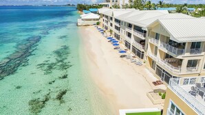 Un hotel frente a la playa con varios balcones, una zona de piscina con sombrillas y aguas turquesas cristalinas.