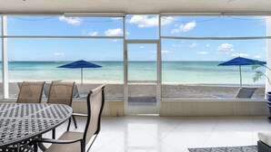 Una habitación con un amplio ventanal que da a una playa, una mesa con sillas y dos sombrillas azules.