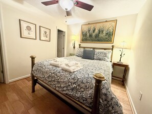 Een slaapkamer met een bed, een plafondventilator, een schilderij en een nachtkastje met een lamp.