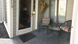 A front porch with a sliding glass door, a wicker basket with flowers, and two wicker chairs with striped cushions.