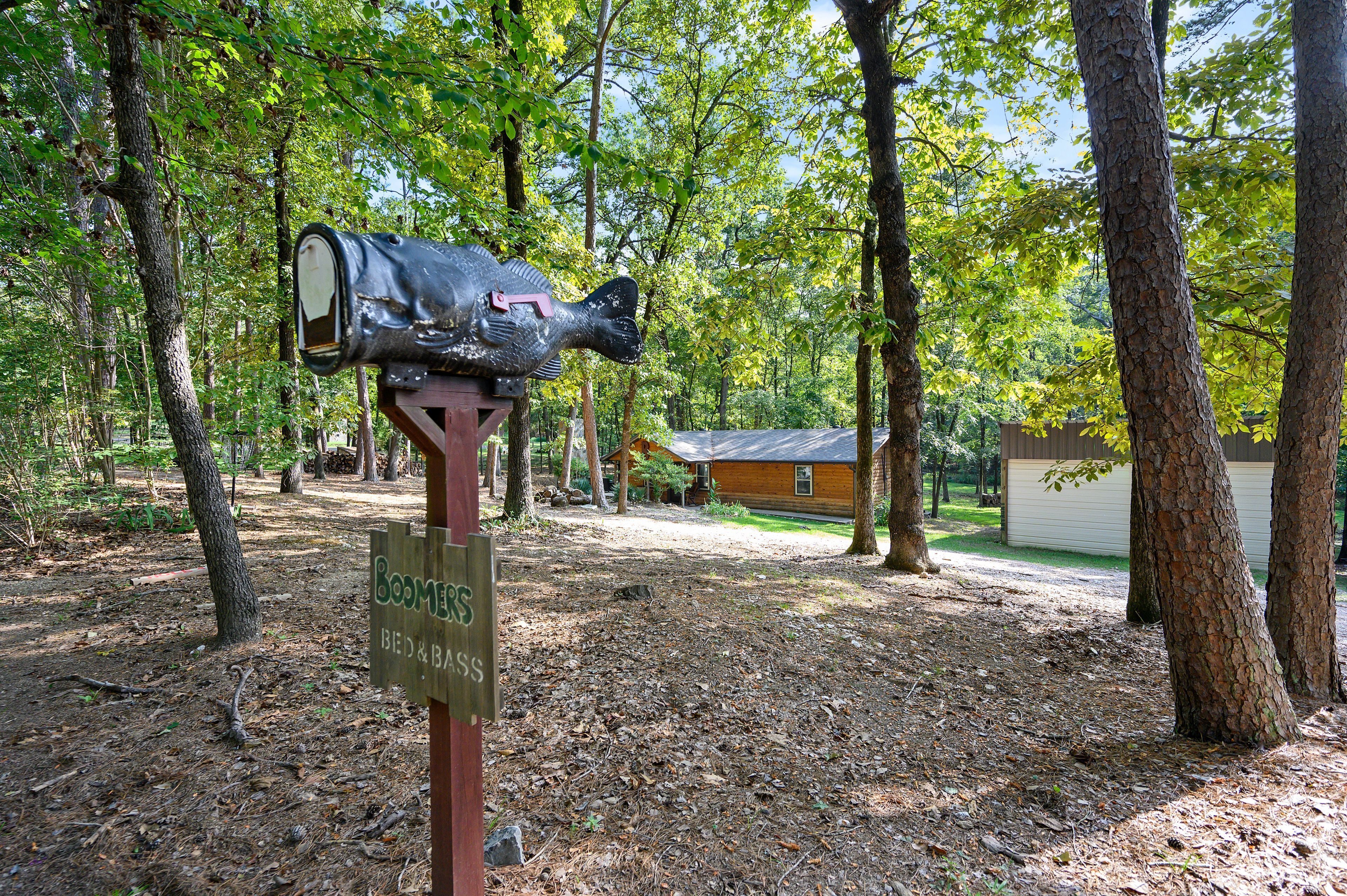 A mailbox with a fish design in a wooded area with a house in the background.