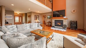 A spacious living room with a fireplace, a large TV mounted above it, and a wooden ceiling. - Luxury Mountain Escape w/ Community Perks (McHenry)