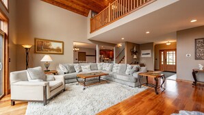 A spacious living room with a high ceiling, wooden flooring, a large sectional sofa, a coffee table, and a staircase leading to an upper level. - Luxury Mountain Escape w/ Community Perks (McHenry)