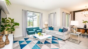 A modern living room with a white sofa, teal armchairs, and a coffee table. There are large windows with curtains, a chandelier, and a dining area visible. - Private Pool + Themed kids rooms! * Near Disney! (Davenport)