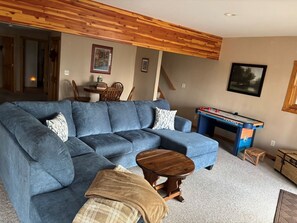 A living room with a blue sectional sofa, a wooden coffee table, a foosball table, and a small wooden stool.