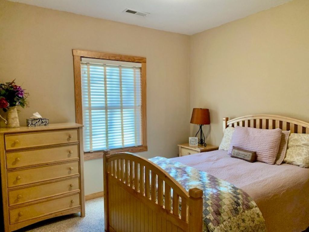 Une chambre à coucher avec un lit en bois, une commode, une table de chevet, une lampe et une fenêtre avec des stores.