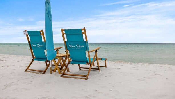 Deux chaises de plage, dotées d’un parasol bleu sur lequel on peut lire « Silver Dunes Destin, Floride », sont installées sur une plage de sable, avec l’océan en arrière-plan.
