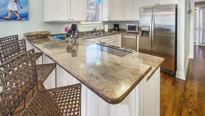 A kitchen with white cabinets, a stainless steel refrigerator, a large island with a sink, and a dining area with wicker chairs. - Beautiful Home at the Front of Seacrest- 3 Bikes (Panama City Beach)