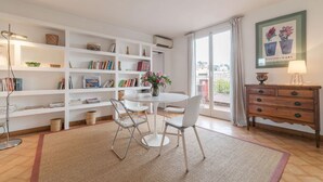 Een helder verlichte kamer met een ronde eettafel, witte stoelen, een boekenkast, een houten dressoir en een groot raam met gordijnen.