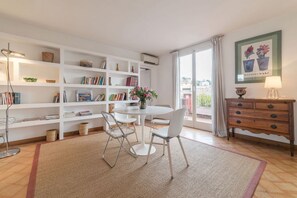A well-lit room with a round dining table, white chairs, a bookshelf, a wooden dresser, and a large window with curtains.