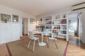 A modern living room with a white dining table, two chairs, a bookshelf, and a small side table with a vase.