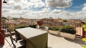 A rooftop terrace with a table and chairs, potted plants, and a view of the cityscape.