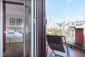 A balcony with a view of a church and buildings, a bed with white linens, and two framed pictures on the wall.